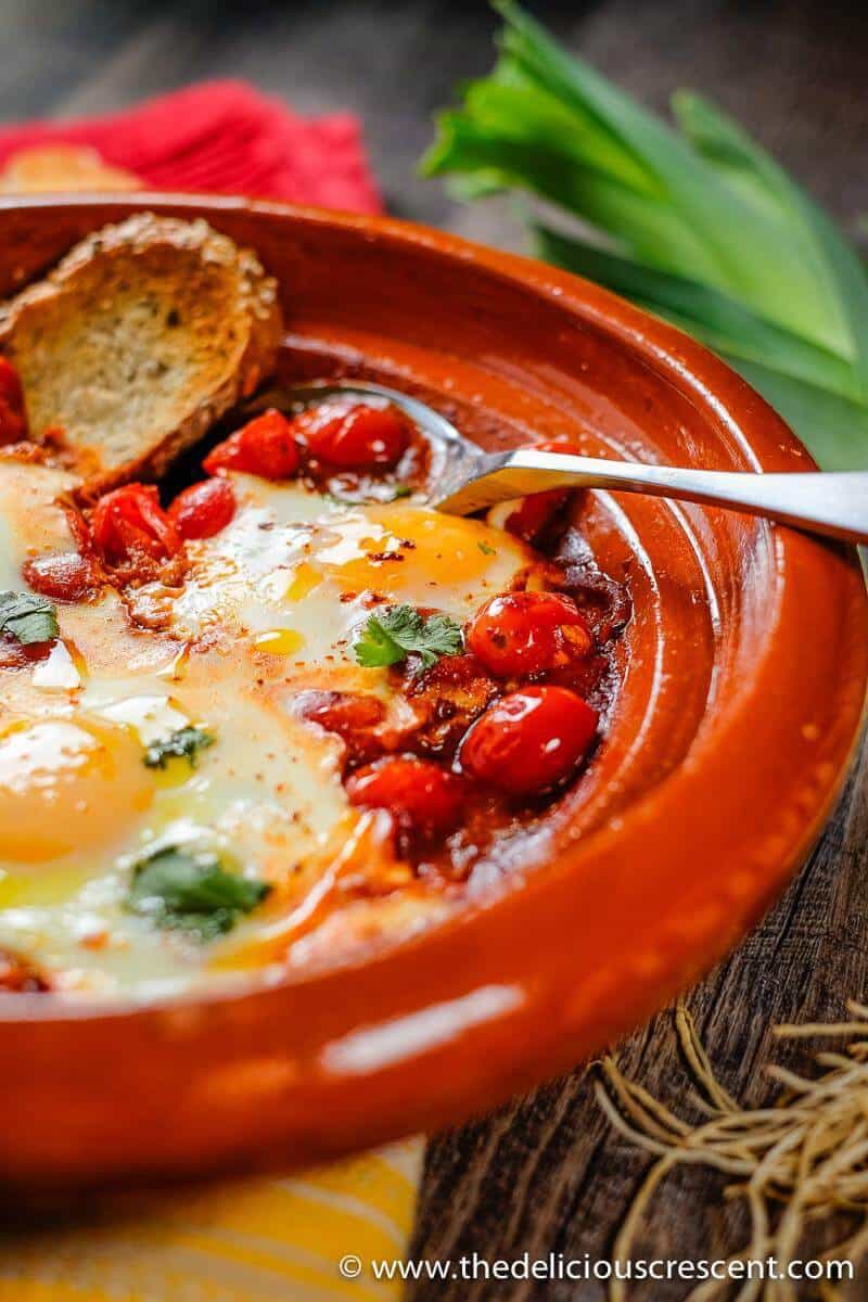 Moroccan eggs in tomato sauce (Shakshuka) is a healthy comfort dish that is so quick and easy to prepare. It is a delicious option for breakfast, brunch or any other meal of the day. This recipe is vegetarian, gluten-free, dairy-free, nut-free and low-carb. #Eggs #Breakfast Moroccan Eggs in Tomato Sauce with a piece of bread and a spoon.