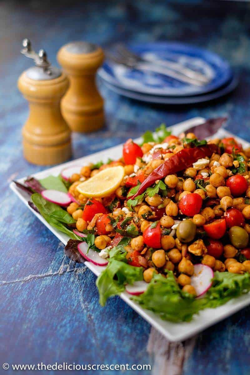 Mediterranean chickpea salad is packed with super delicious flavors and textures. It is an easy, meatless salad that is filling, healthy and packed with plant protein and fiber. Make this recipe vegan by skipping feta cheese. This simple gluten free salad with the spicy dressing will make a great meal. #chickpeasalad #Mediterraneanfood #middleeasternfood A side view of the chickpea salad on a white serving plate.