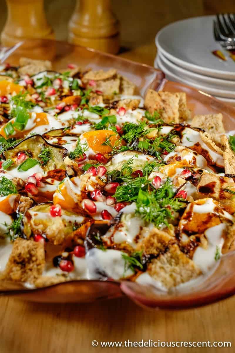 Eggplant yogurt salad is an adapted version of the popular middle eastern dish “Fatteh” with added butternut squash. It is a savory salad with roasted vegetables in creamy garlic yogurt sauce, topped with pomegranate, walnuts and crispy pita chips and often served in a casserole dish. This Lebanese and Syrian vegetarian recipe is heart healthy and a source of probiotics. #mediterraneandiet #sidedish Eggplant fatteh topped with creamy yogurt sauce, pomegranate, walnuts and crispy pita chips.