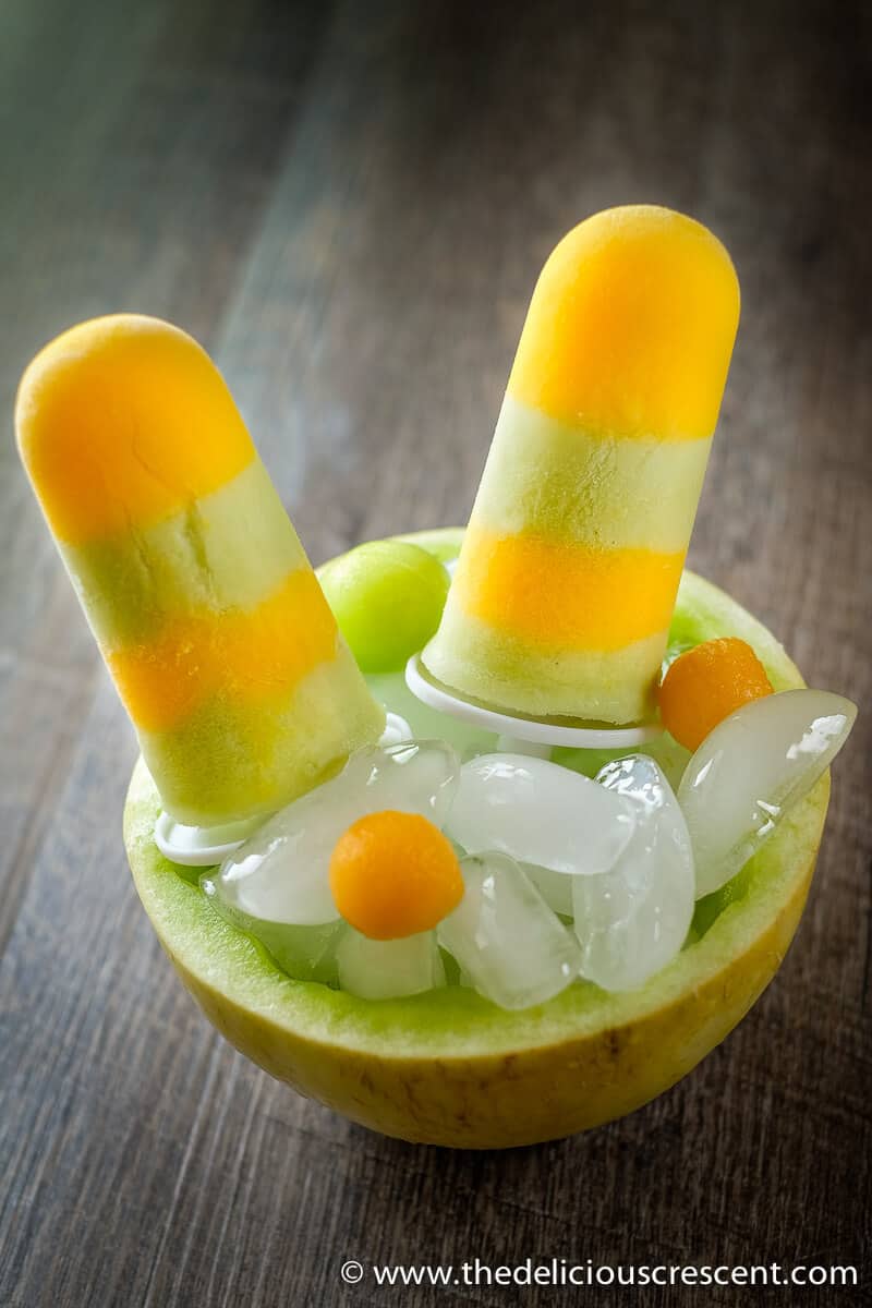 Two melon popsicles inserted in a stack of ice cubes inside a hollowed out melon bowl.