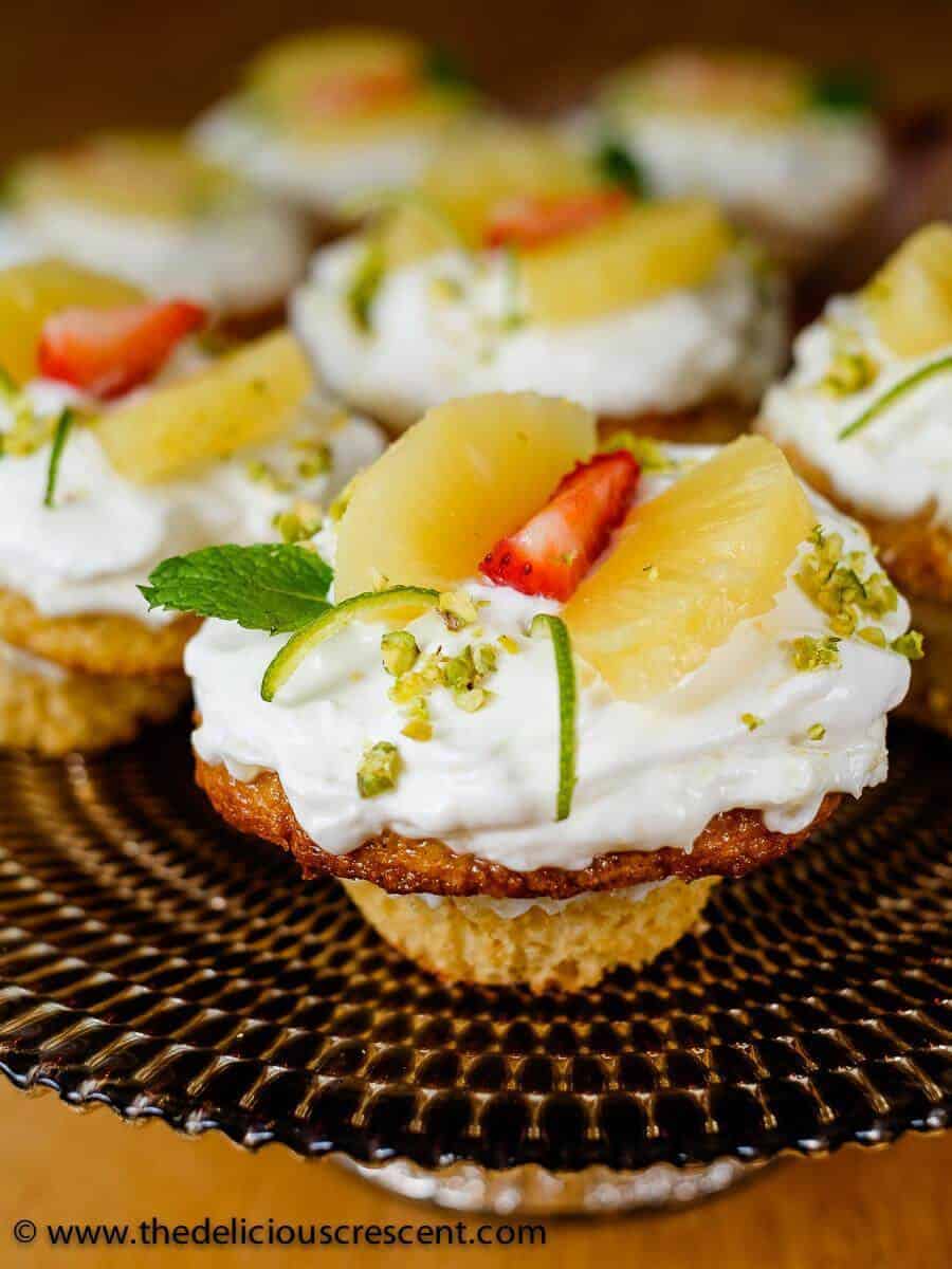 Pineapple cupcakes made with fresh cream and yogurt are amazingly delicious. These cute homemade cupcakes have an easy decoration concept and would be a great party idea for kids. This recipe made from scratch allows you to use thick Greek style yogurt to lighten up a traditional favorite, enhancing the nutrition, and is still absolutely delicious. Try this sweet treat this holiday season. #cupcakes #pineapplecake Several pineapple cupcakes arranged on a brown plate.