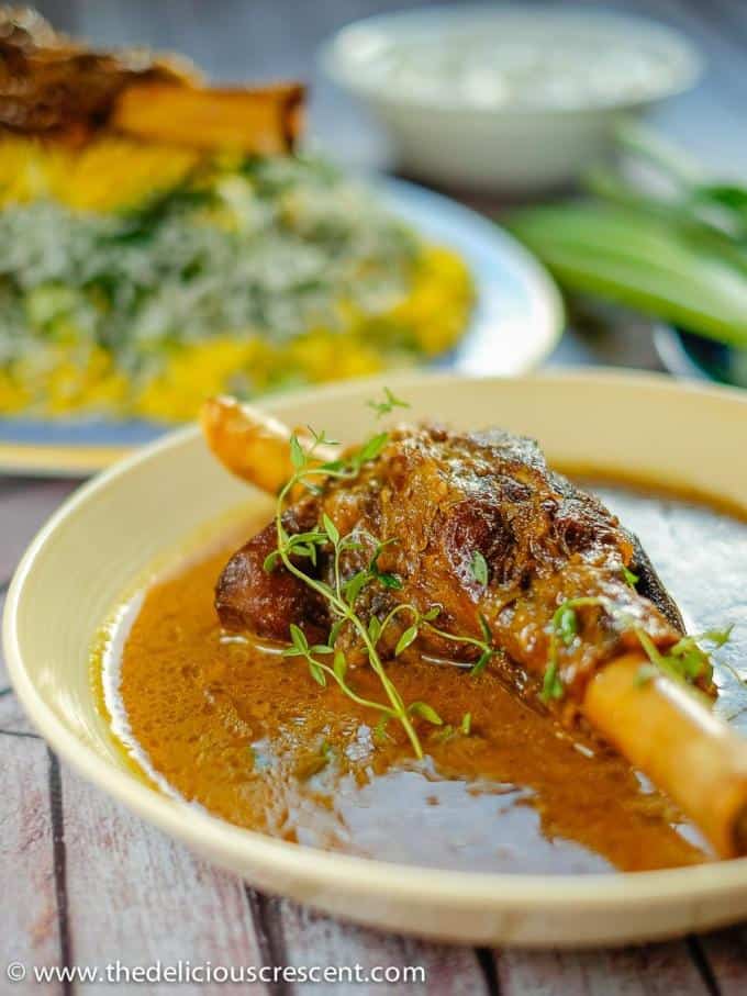 Braised lamb shanks slow cooked in an aromatic sauce with Persian spices, saffron, lemon juice and herbs makes a spectacular feast. The braising technique will give you succulent lamb with fall-off-the-bone tender meat in an amazingly delicious sauce! Let me show you how to cook lamb shanks in cast iron skillet or dutch oven with this easy middle eastern recipe. Including tips for cooking in oven or crockpot. #lambshanks #Persianfood Lamb shanks braised in an amazing Persian sauce and served in a round dish with rice and yogurt salad.