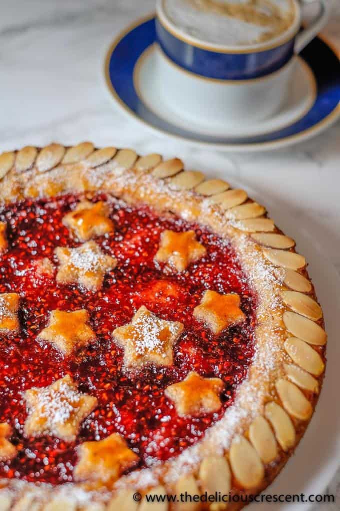 Linzer torte is a nutty, crumbly, rich pastry filled with jam and is as enchantingly pretty as it is tasty. It is easier than you would expect and delicious to the last bite! Perfect for entertaining. This dessert recipe is a specialty of Germany, Austria etc and is filled with raspberry jam and popular for holiday baking. Including suggestions for gluten free and dairy free options. #holidaybaking #torte #dessert. Linzer torte decorated with star shapes and placed on a table.