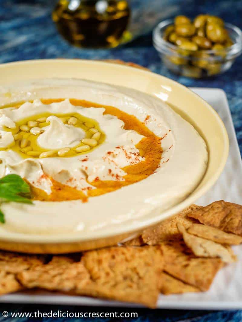 Homemade hummus served in a cream bowl along with pita chips and green olives.